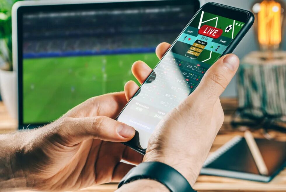 Mãos segurando celular com aplicativo de apostas ao vivo durante partida de futebol
